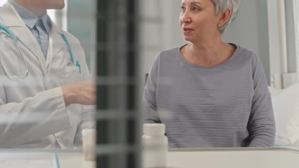 rajattu laukaus mies lääkäri valkoinen lab takki antaa pillereitä vanhempi aasialainen nainen istuu sängyllä kirkas sairaala osasto - Materiaali, video