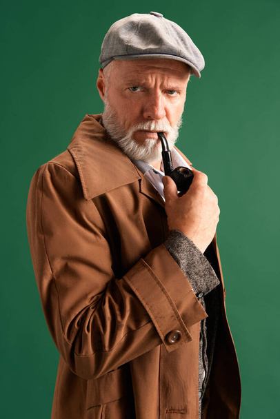 Portret van een zelfverzekerde en strikte man, detective met vintage hoed, elegant pak met pijp in de mond kijkend naar camera op groene achtergrond. Concept van mode, emoties, retro. Steun. - Foto, afbeelding