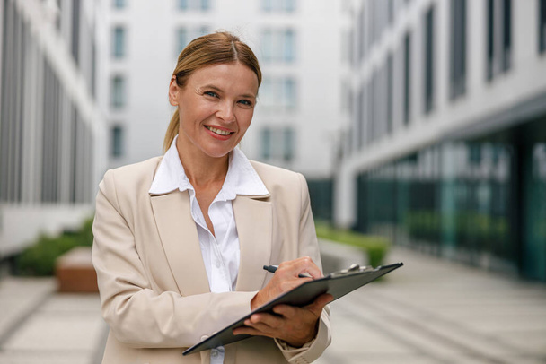 Улыбающаяся женщина-офисный работник стоит на современном фоне здания и делает заметки - Фото, изображение