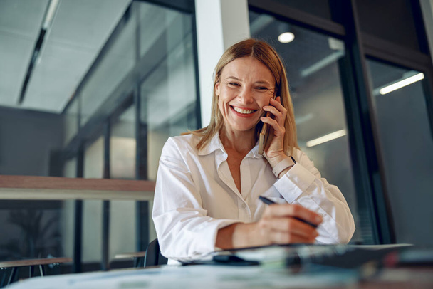居心地の良いオフィスに座って電話で話している間,ラップトップで働く笑顔の成熟したビジネス女性  - 写真・画像