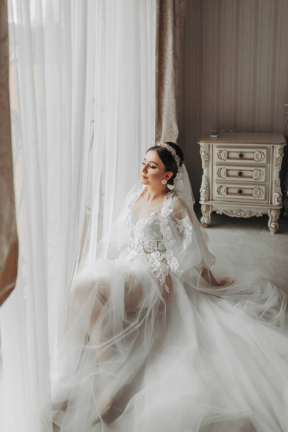 a beautiful girl with a wedding hairstyle and a tiara on her head in a transparent robe is preparing for a wedding in a hotel with a royal interior. Long veil, natural light, portrait photo - 写真・画像