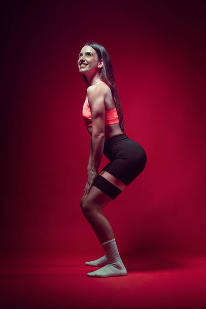 Junge sportlich attraktive kaukasische Sportlerin posiert in rosa Tank-Top und schwarzem Sauna-Shorts-Fitness-Outfit. Studioshooting mit hübscher schlanker Brünette beim Aerobic Stretching mit Gummibändern - Foto, Bild