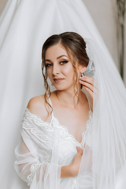 Morning of the bride before the wedding. A beautiful young woman in a white robe near her dress. Natural beauty and professional makeup - Fotografie, Obrázek