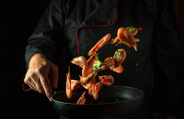 El cocinero en su mano sostiene una sartén con camarones congelados y perejil. Aislado sobre fondo negro. Recetas de mariscos y mariscos y cocina casera. - Foto, Imagen
