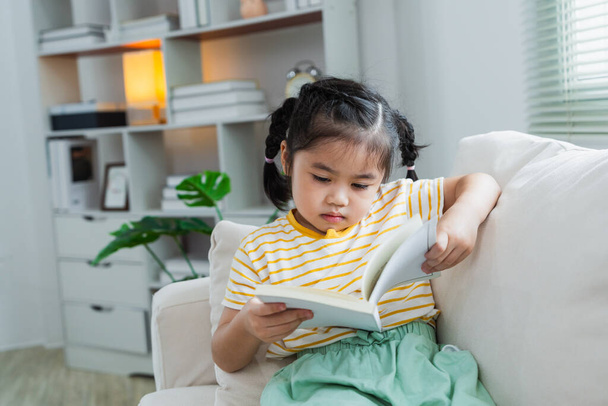 Χαρούμενη ευτυχισμένη ασιατική κοριτσάκι κοριτσάκι χαμογελώντας και διαβάζοντας το βιβλίο, ενώ κάθεται στον καναπέ καναπέ στο σαλόνι στο σπίτι. Κορίτσι relex ανάγνωση βιβλίο χαμόγελο στον καναπέ στο σπίτι. Επιστροφή στο σχολείο έννοια. - Φωτογραφία, εικόνα