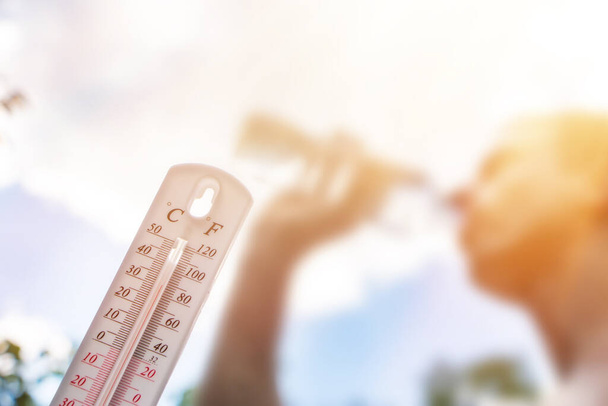 Άνθρωπος πόσιμο νερό σε ακραία θερμότητα, θερμόμετρο κατά τη διάρκεια της καλοκαιρινής ημέρας δείχνει ή δείχνουν υψηλό βαθμό θερμοκρασίας με ήλιο στο παρασκήνιο. - Φωτογραφία, εικόνα