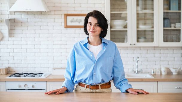 Mladá žena stojící v blízkosti jídelního stolu na pozadí kuchyně a při pohledu na kameru, připravuje vařit zdravé snídaně a nahrávat video pro kulinářský blog. - Fotografie, Obrázek