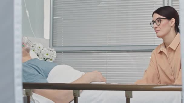 Młoda biała kobieta odwiedza chorego męża w szpitalu, leżącego na łóżku w masce tlenowej - Materiał filmowy, wideo