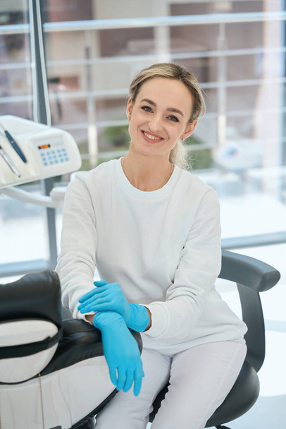 Молодая женщина в защитных перчатках сидит на стуле, готовится к работе в стоматологии - Фото, изображение