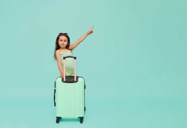 Белая симпатичная маленькая девочка-путешественница в летней одежде, позирует с бирюзовым чемоданом, отводит в сторону рекламное пространство на изолированном голубом фоне студии, уверенно смотрит в камеру. Дети и путешествия - Фото, изображение