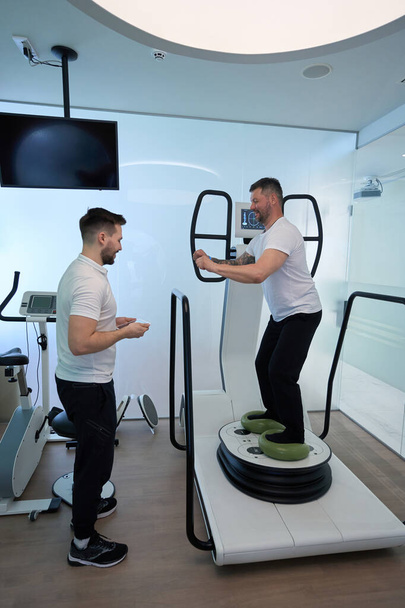 Мужчина делает упражнения на равновесии на многоосной моторизованной платформе в присутствии физиатра - Фото, изображение