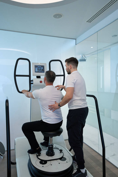 Adult person working out in seated position on multi-axis motorized platform with assistance of physiotherapist - Foto, afbeelding