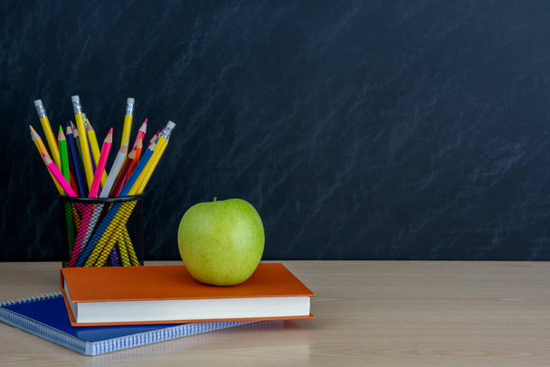 metalen houder met kleurpotloden en groene appel en boeken op bureau op zwarte schoolbordachtergrond - Foto, afbeelding