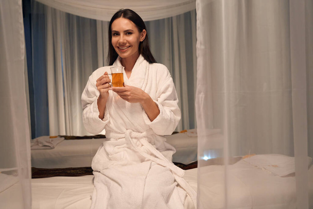 Joyous spa client in bathrobe seated on massage couch holding cup of beverage in her hands - Photo, Image