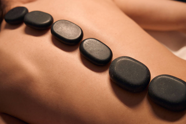 Closeup of naked woman lying prone with flat black stones positioned along spine - Photo, Image