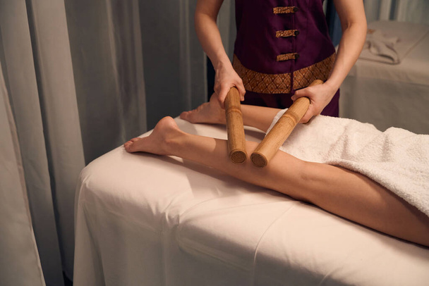 Vystřižená fotka masoterapeuta válcování dvojice bambusových tyčí přes nohu klienta během masoterapie - Fotografie, Obrázek