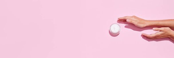 Bovenaanzicht van jonge tedere vrouwen handen houden open potten cosmetische crèmes op roze achtergrond. Vrouwelijke gezichtsverzorging. Concept van lichaams- en huidverzorging, spa, dermatologie, behandelingen, anti-aging. Banner voor reclame - Foto, afbeelding