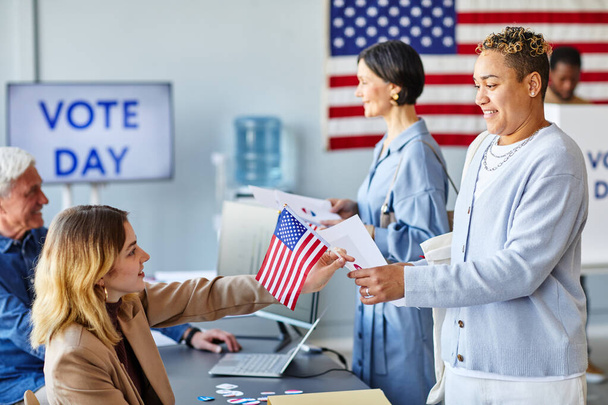 Retrato de vista lateral de una sonriente mujer negra que vota en el centro de votación con bandera estadounidense - Foto, imagen