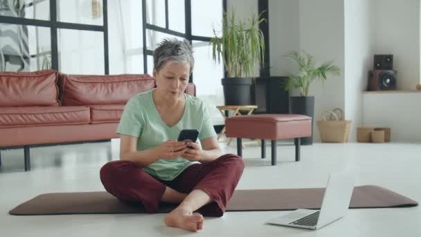 Vieille femme faire des exercices de yoga à la maison. Femme mûre Utilisez le téléphone mobile pour la navigation en ligne ou les achats sur Internet. Les femmes utilisent un smartphone. Images 4k de haute qualité - Séquence, vidéo