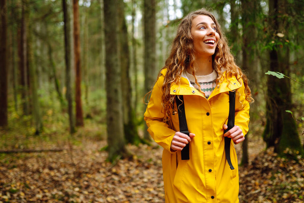 Female tourist in a yellow coat with a backpack traveling along a wooden path in the forest. Trekking trail. Concept of travel, nature, vacation. - Фото, изображение