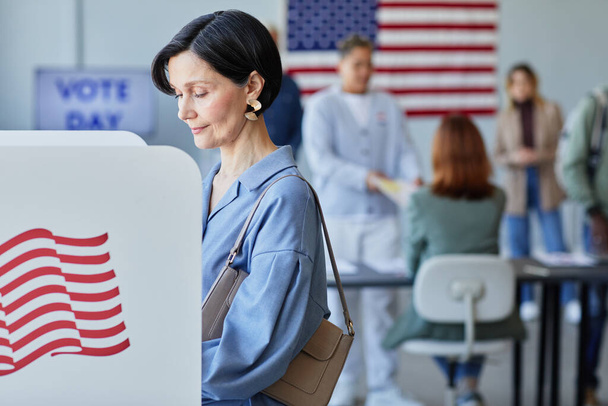 Seitenansicht Porträt einer eleganten erwachsenen Frau, die am Wahltag in der Wahlkabine ihre Stimme abgibt, mit amerikanischen Flaggen in Schuss, Kopierraum - Foto, Bild