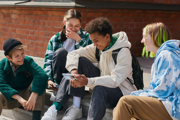 Grupo de amigos sentados em passos de construção ao ar livre e rindo enquanto assiste vídeo engraçado no telefone móvel - Foto, Imagem