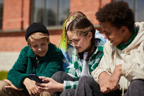 Adolescente en lunettes communiquant en ligne sur téléphone mobile tout en étant assise à l'extérieur avec ses amis - Photo, image