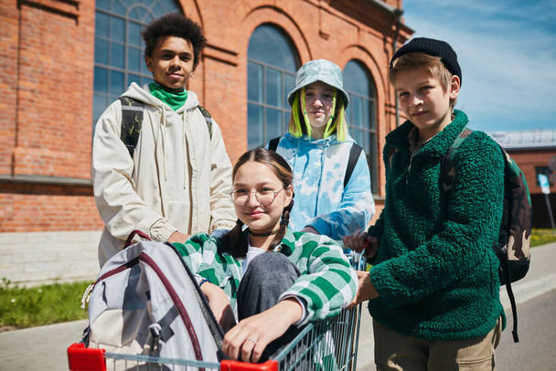 Portrét skupiny dospívajících chlapců a dívek při pohledu na kameru při jízdě na nákupním košíku na ulici - Fotografie, Obrázek