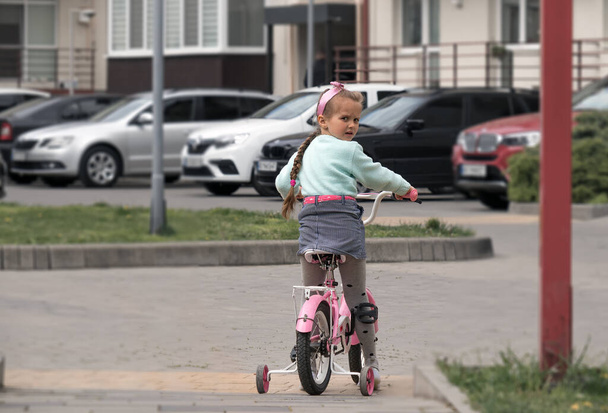 pequena menina europeia, com um longo pigtail, 5-6 anos, monta uma bicicleta ao longo da rua da cidade, um passeio de bicicleta infantil - Foto, Imagem