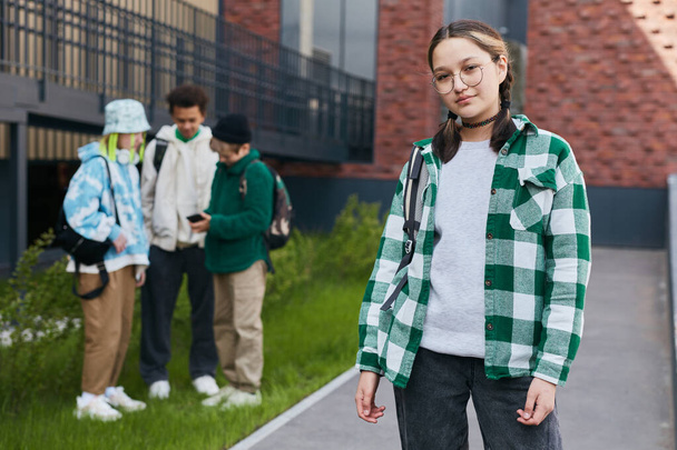 Retrato de adolescente en gafas mirando a la cámara de pie en el patio de la escuela con sus amigos en el fondo - Foto, Imagen