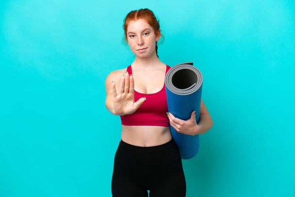 Jeune femme rougeâtre sportive allant à des cours de yoga tout en tenant un tapis isolé sur fond bleu faisant geste d'arrêt - Photo, image