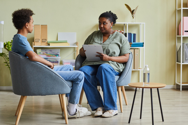 Afrikanischer Schulpsychologe spricht während der Sitzung mit schwierigen Teenagern, während sie im Büro auf Sesseln sitzen - Foto, Bild