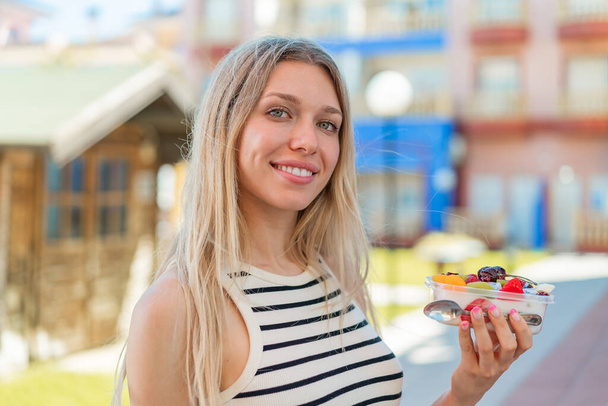 Mujer rubia joven sosteniendo un tazón de fruta al aire libre sonriendo mucho - Foto, Imagen