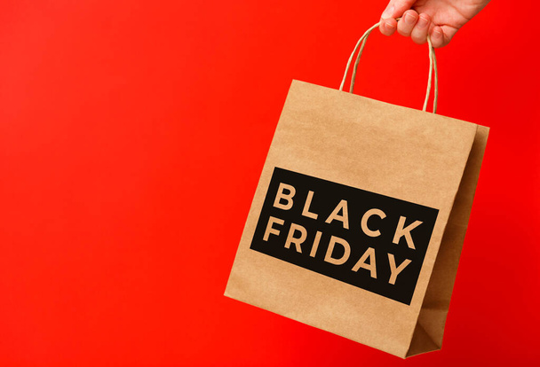 Female hand holding craft shopping bag with Black Friday text on red background. Sale, discount, shopping concept - Photo, Image