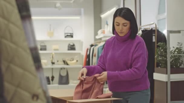 Nuori nainen kauppa-avustaja ottaa design-vaatteita pahvilaatikosta ja ripustaa ne ripustimeen työskennellessään putiikissa - Materiaali, video