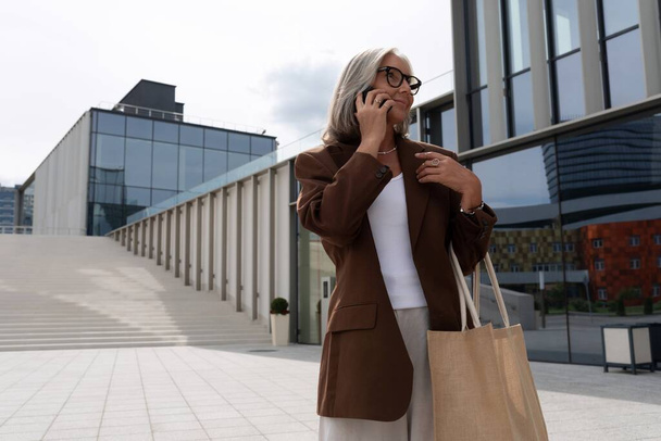 een grijs-harige vrouw van middelbare leeftijd met een bob kapsel elegant gekleed loopt rond het business center op straat. - Foto, afbeelding