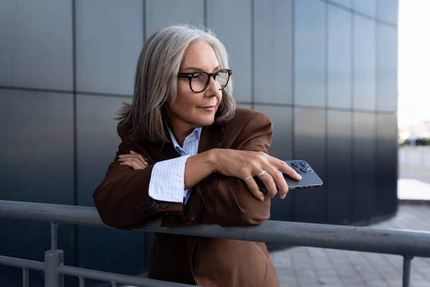 60 años de edad exitosa esbelta mujer de negocios de pelo gris vestida con una elegante chaqueta marrón está descansando de pie sobre el telón de fondo de una pared del edificio. - Foto, Imagen