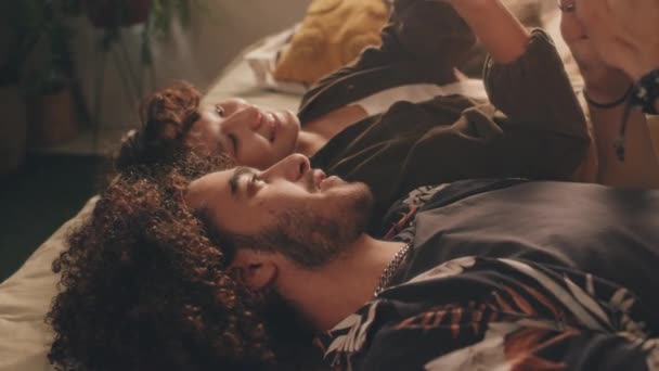 Pevná střední záběry mladého muže a mladé dívky, oba kudrnatí, ležící v posteli s telefony, ukazující si obrazovky a usmívající se. Okno a rostlina v pozadí, sluneční světlo, boční pohled - Záběry, video