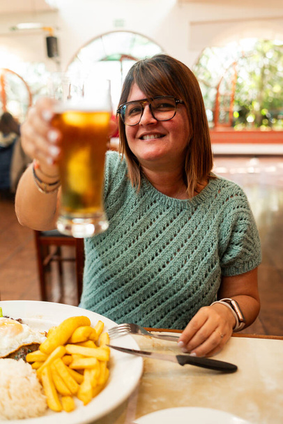 vertikales Bild einer Frau, die mit einem Bier in der Hand in einer Bar feiert, in der Essen serviert wird. - Foto, Bild