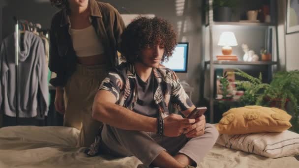 Full medium shot of curly young man sitting on his bed with phone while young girl coming to him from behind, hugging, putting his phone away, both smiling. Clothes, monitor, lamp, plant in background - Footage, Video