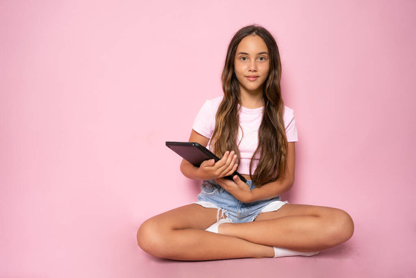 Nuori tyttö istuu lattialla ja käyttää tabletin eristetty vaaleanpunainen tausta. - Valokuva, kuva