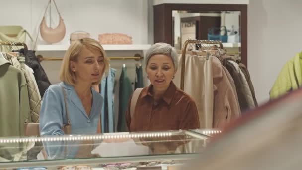 Dvě elegantní moderní ženy při pohledu na skleněný pult displej a diskutovat o položkách při nakupování v drahé značkové butik - Záběry, video