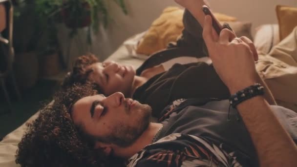 Loose close-up of young man and young girl, both curly, lying in bed with their phones, looking at each other screens, both smiling. High angle view - Footage, Video