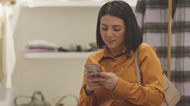Joven hermosa mujer de Oriente Medio hablando en el teléfono inteligente mientras compra ropa en boutique de diseño - Metraje, vídeo