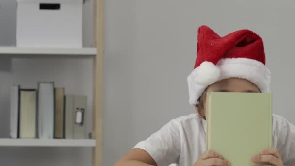 Junge von 9 Jahren in Santas Hut versteckt sich hinter dem Buch. Nahaufnahme. Hochwertiges 4k Filmmaterial - Filmmaterial, Video