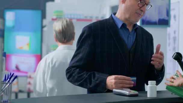Старый клиент прибывает в аптеку кассу после того, как находит необходимые медицинские товары, покупает таблетки от боли в животе с помощью кредитной карты, ему помогает фармацевт с сумкой - Кадры, видео
