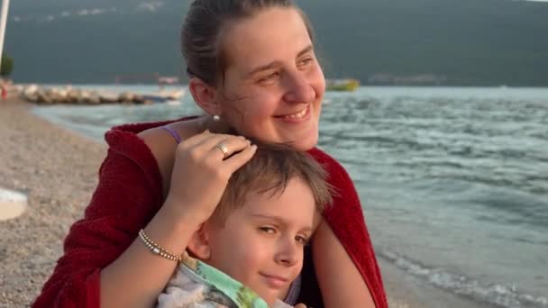 Молодая любящая мать обнимает своего маленького сына, укрываясь пляжным полотенцем и чувствуя холод в море. Отдых, летний отдых и туризм. - Кадры, видео