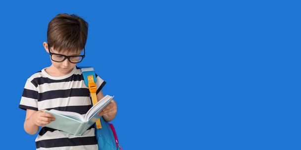 Mavi arka planda kitabı ve sırt çantası olan küçük bir okul çocuğu. - Fotoğraf, Görsel