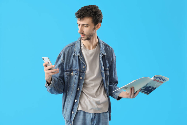 Όμορφος νεαρός άνδρας με κινητό τηλέφωνο και περιοδικό σε μπλε φόντο - Φωτογραφία, εικόνα