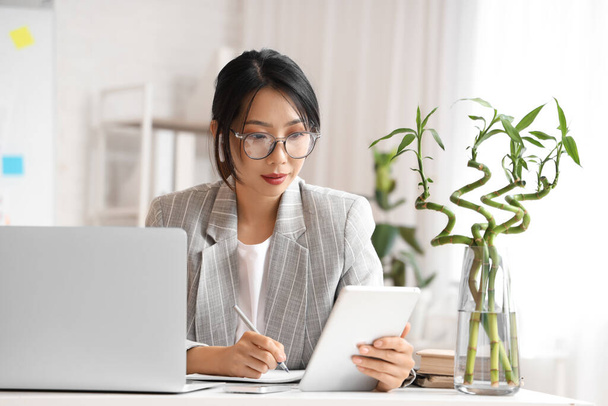 Jovem empresária asiática sentada à mesa e trabalhando com laptop e tablet no escritório - Foto, Imagem